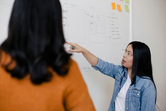 Two people stand by a whiteboard, one person points to the board